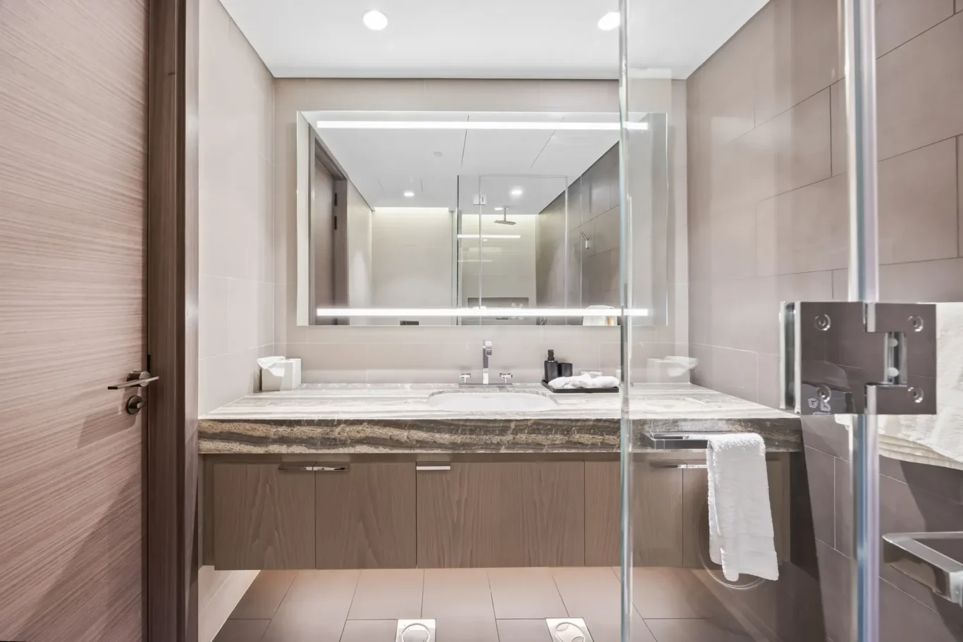 Modern bathroom featuring a wide marble countertop, undermount sink, and wooden vanity cabinets beneath; a large backlit mirror spans the wall and a glass shower enclosure is on the right.