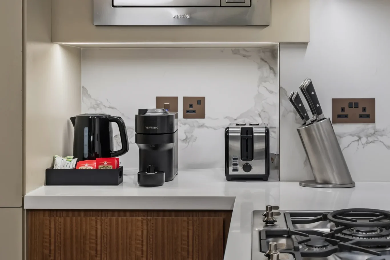 Countertop coffee station with kettle, Nespresso machine, toaster, knife block, and tea sachets against a marble backsplash under-soft lighting.