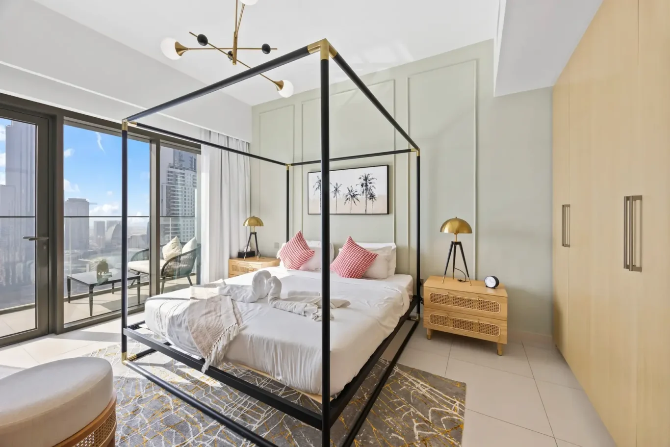 Modern bedroom with a black metal four-poster bed, palm artwork, and city view through floor-to-ceiling glass doors on a balcony."],