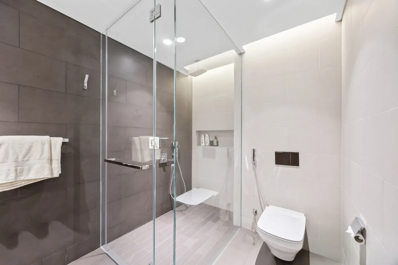 Modern bathroom with a glass shower enclosure, dark brown wall tiles, light gray floor tiles, towel rack, and a wall-hung toilet.