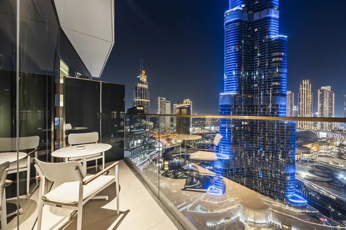 Nighttime balcony seating with white chairs and round tables overlooking a brightly lit city skyline, including a tall blue-lit skyscraper.