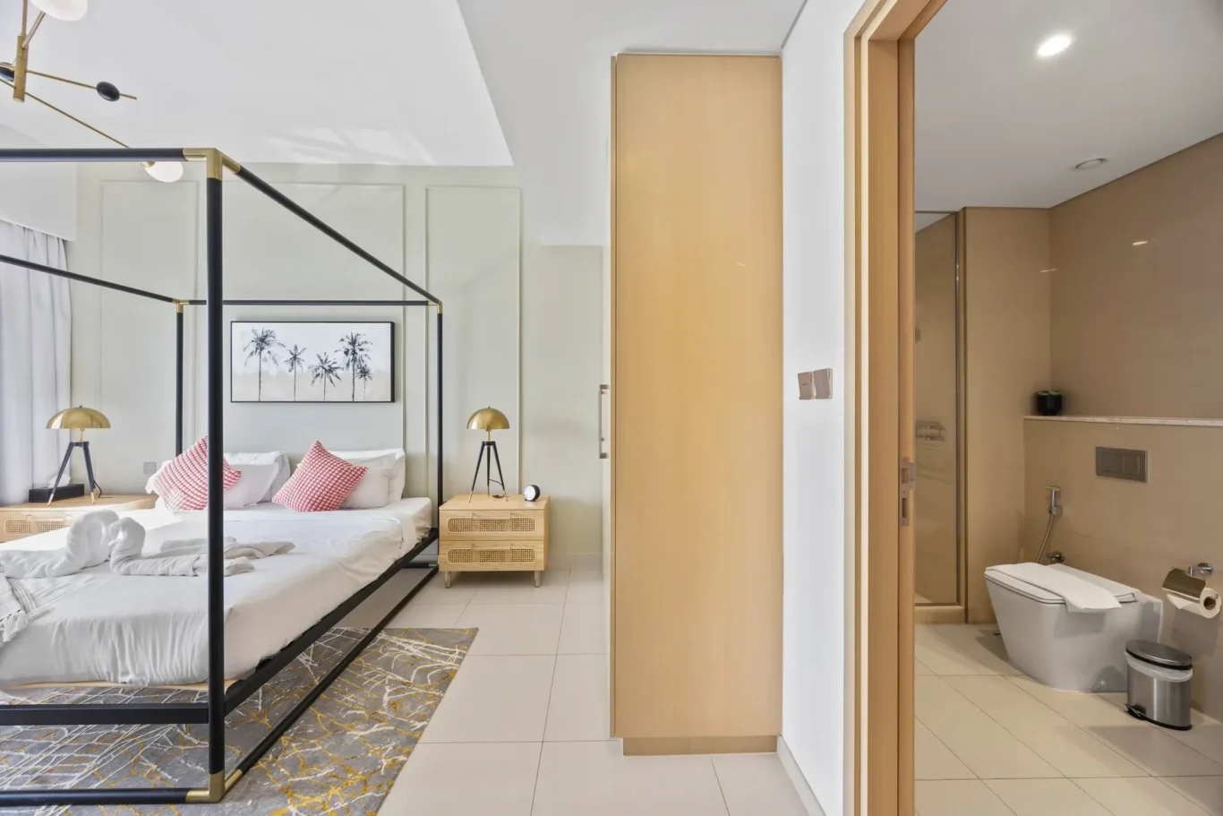 Hotel bedroom with a black metal four‑poster bed, white bedding, and palm artwork; ensuite bathroom visible through a glass door on the right.