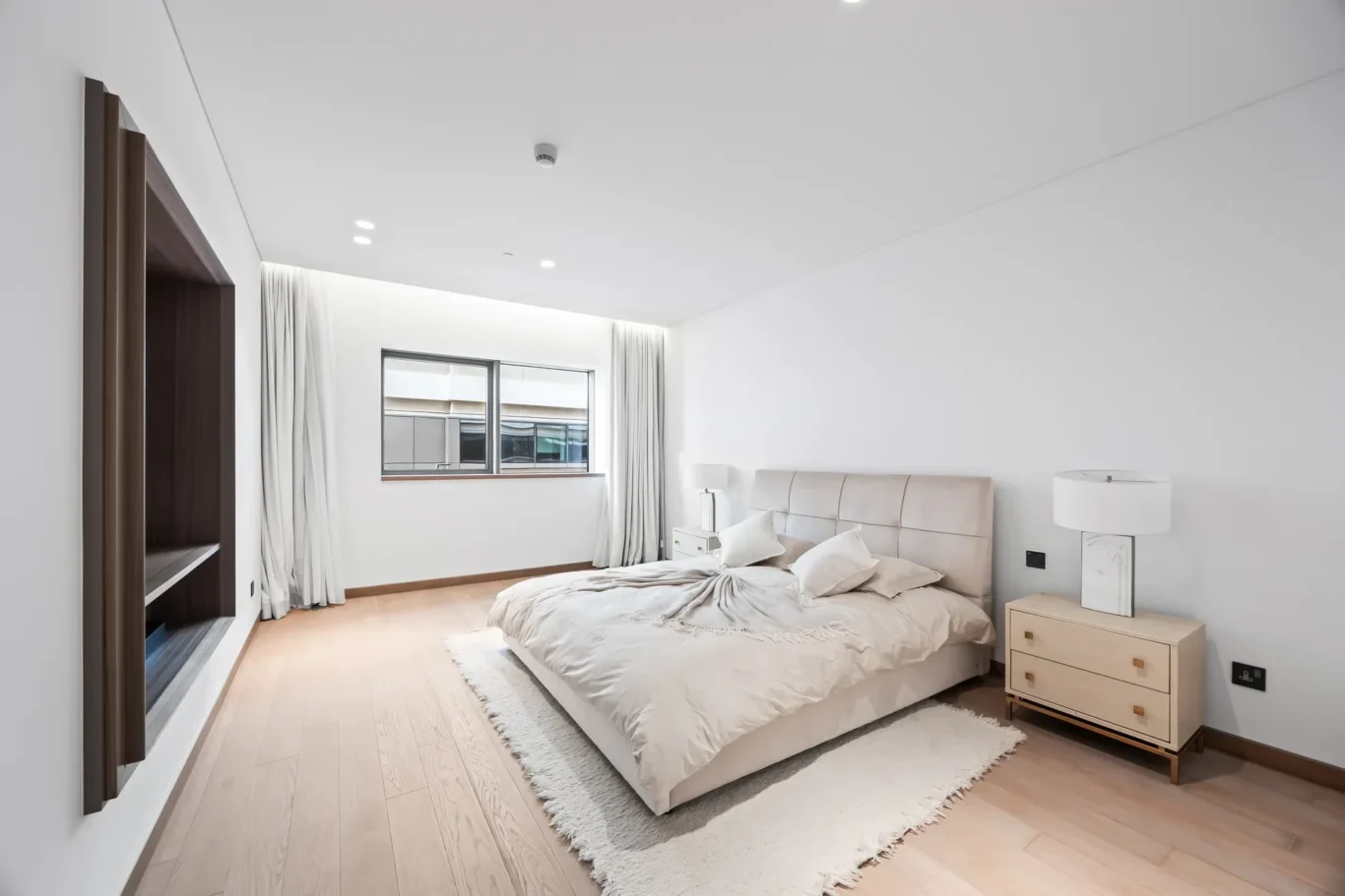 Minimalist modern bedroom with a beige upholstered bed, two nightstands, and white lamps on a light wood floor.