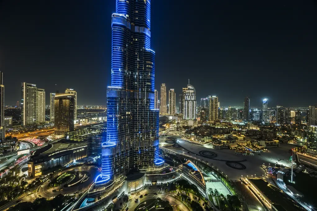 Night skyline of Dubai featuring a tall blue-lit skyscraper surrounded by illuminated buildings and roads at night.