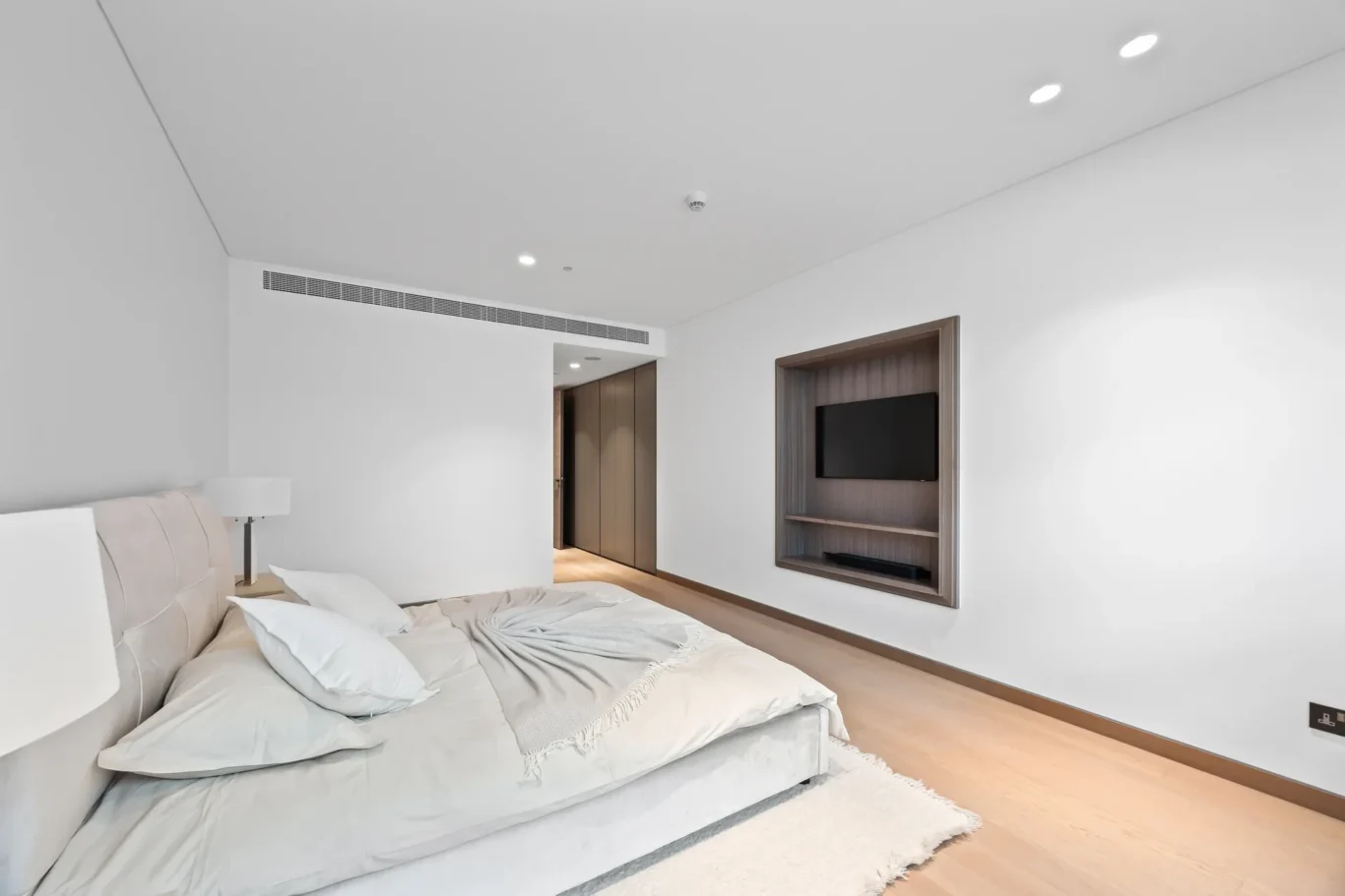 Modern bedroom with a low upholstered bed, pale bedding, bedside lamp, and a wall-mounted TV set into a wood-framed alcove opposite.