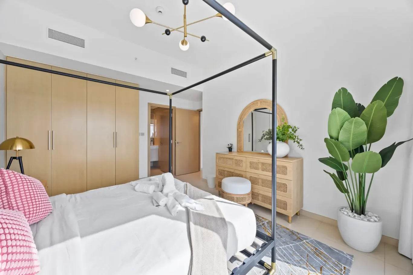 Bright contemporary bedroom with a black four-poster bed, light wood wardrobes, and a woven dresser with a round mirror.