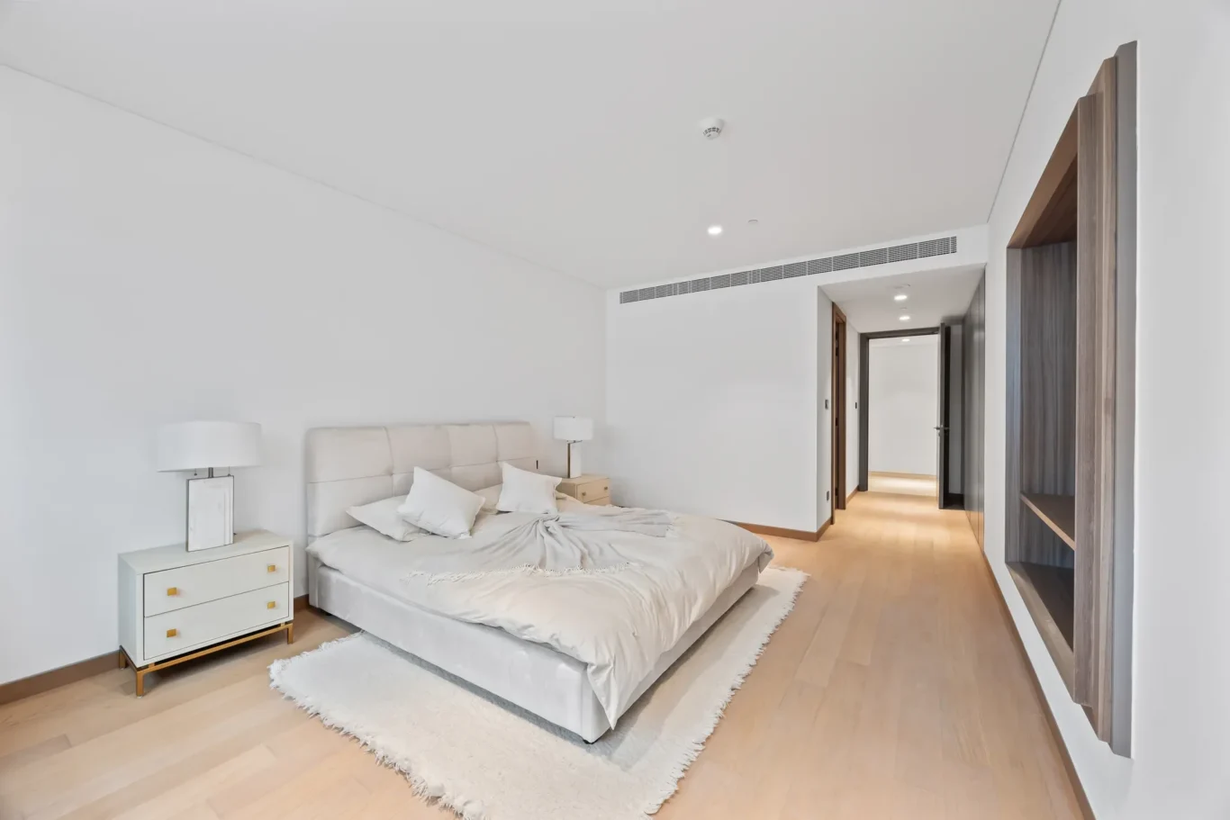Bright minimalist bedroom with a low gray upholstered bed, white bedding, and matching nightstands with lamps; hallway visible in the background in a modern home.