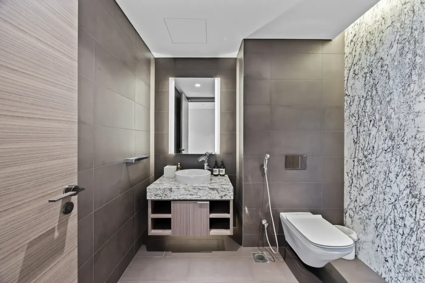 Modern bathroom with gray tiled walls, marble countertop vanity, and a wall-mounted faucet with a round vessel sink. A backlit rectangular mirror, open shelves, and a marble accent wall to the right visible above a wall-mounted toilet with handheld bidet.