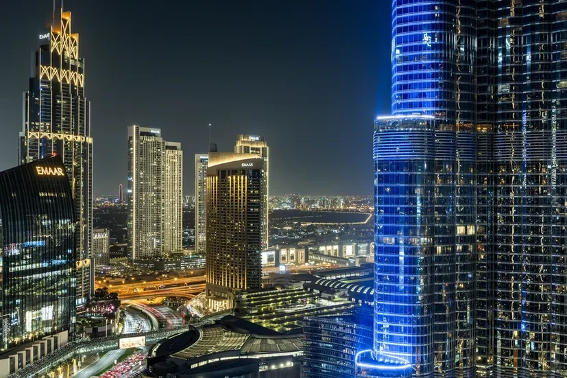 Nighttime city skyline with illuminated skyscrapers, including a tall blue-lit glass tower on the right.