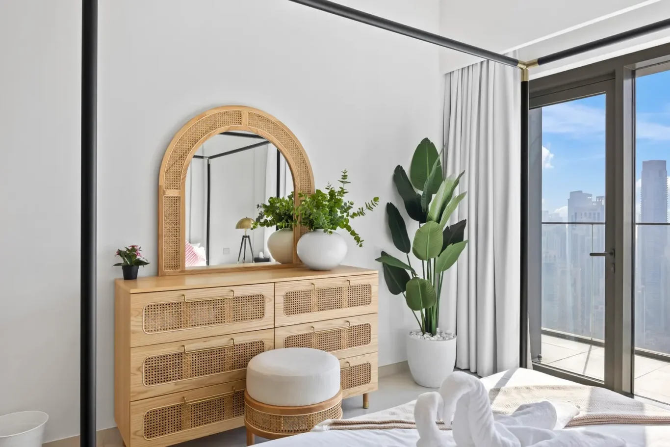 Wooden dresser with woven rattan drawers and an arched mirror, flanked by potted plants in a bright bedroom with a city view through glass doors.