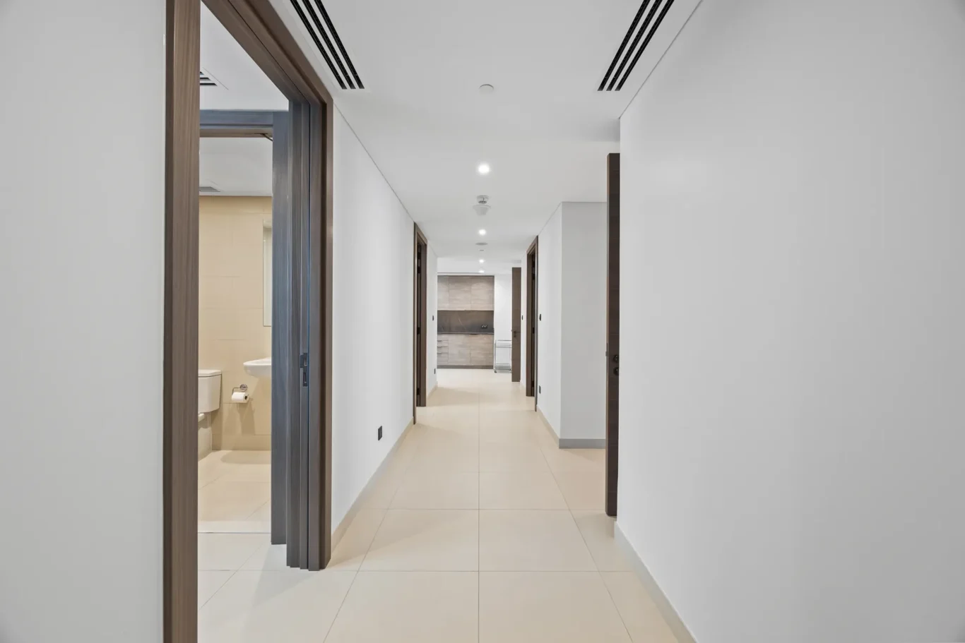 Long modern hallway with beige tiled floor and white walls, and dark wood door frames leading to a kitchen at the far end, with doors on both sides.