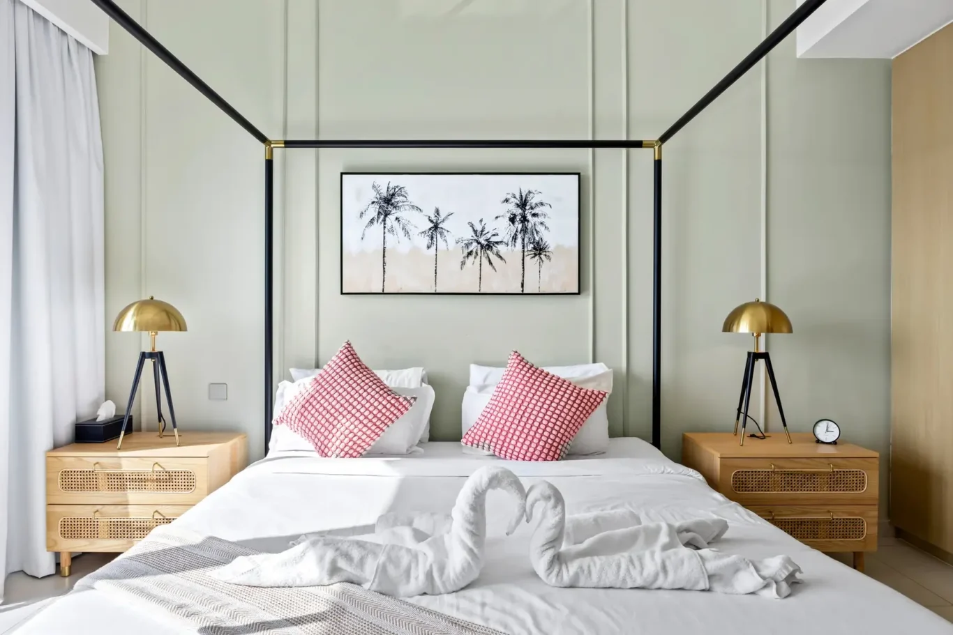 Hotel-style bedroom with a black metal canopy bed, palm tree artwork, white bedding, and two wooden nightstands with gold lamps on each side.
