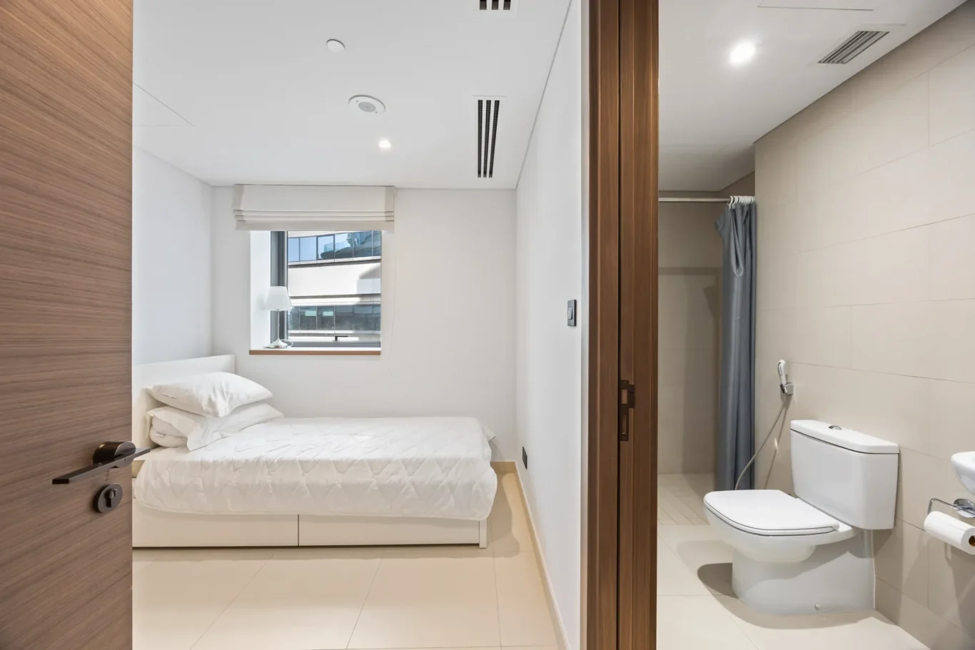 Contemporary hotel bedroom with white bed, pillows, and a window; doorway reveals a tiled bathroom with a toilet and shower curtain.