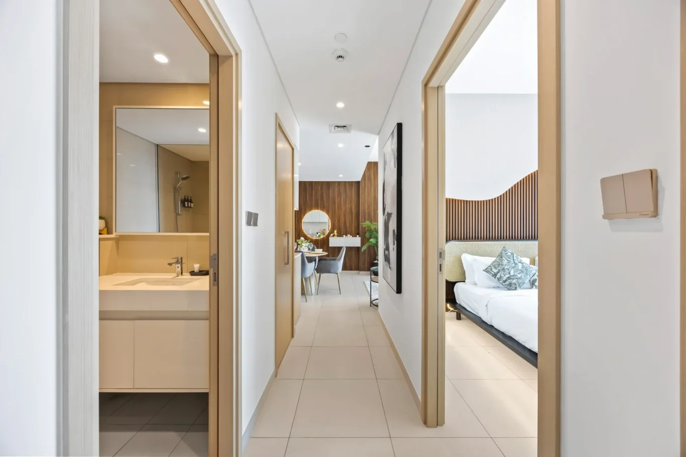 Hallway of a modern hotel suite with a bathroom door on the left and a bedroom on the right featuring a wooden headboard.