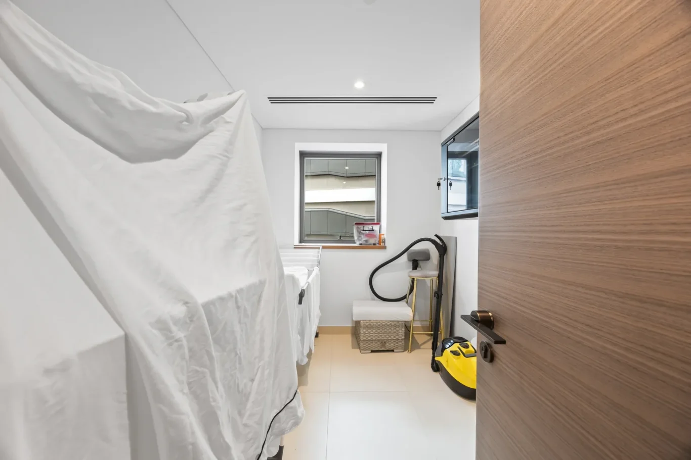 Narrow laundry room with two white-covered machines on the left, a window at the back, and a wooden door on the right.