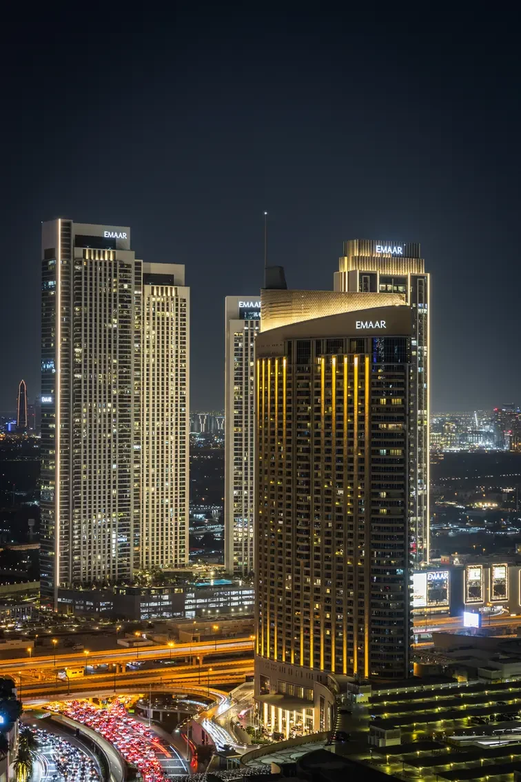 Nighttime cityscape showing illuminated Emaar skyscrapers and a curving, busy highway below with streaks of red and white lights.