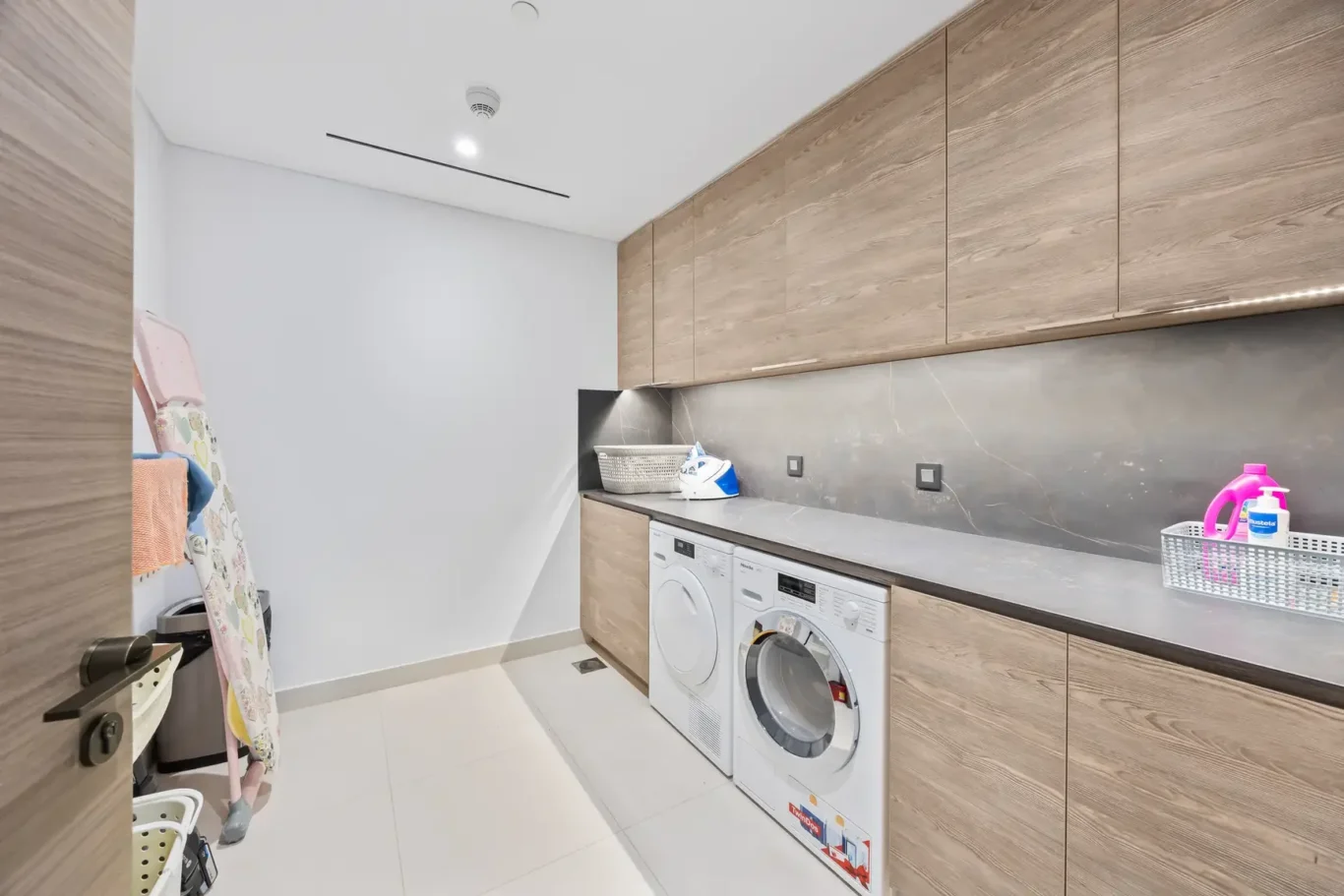 Laundry room with front-loading washer and dryer under a wooden cabinet run, gray marble countertop, and white-tile floor