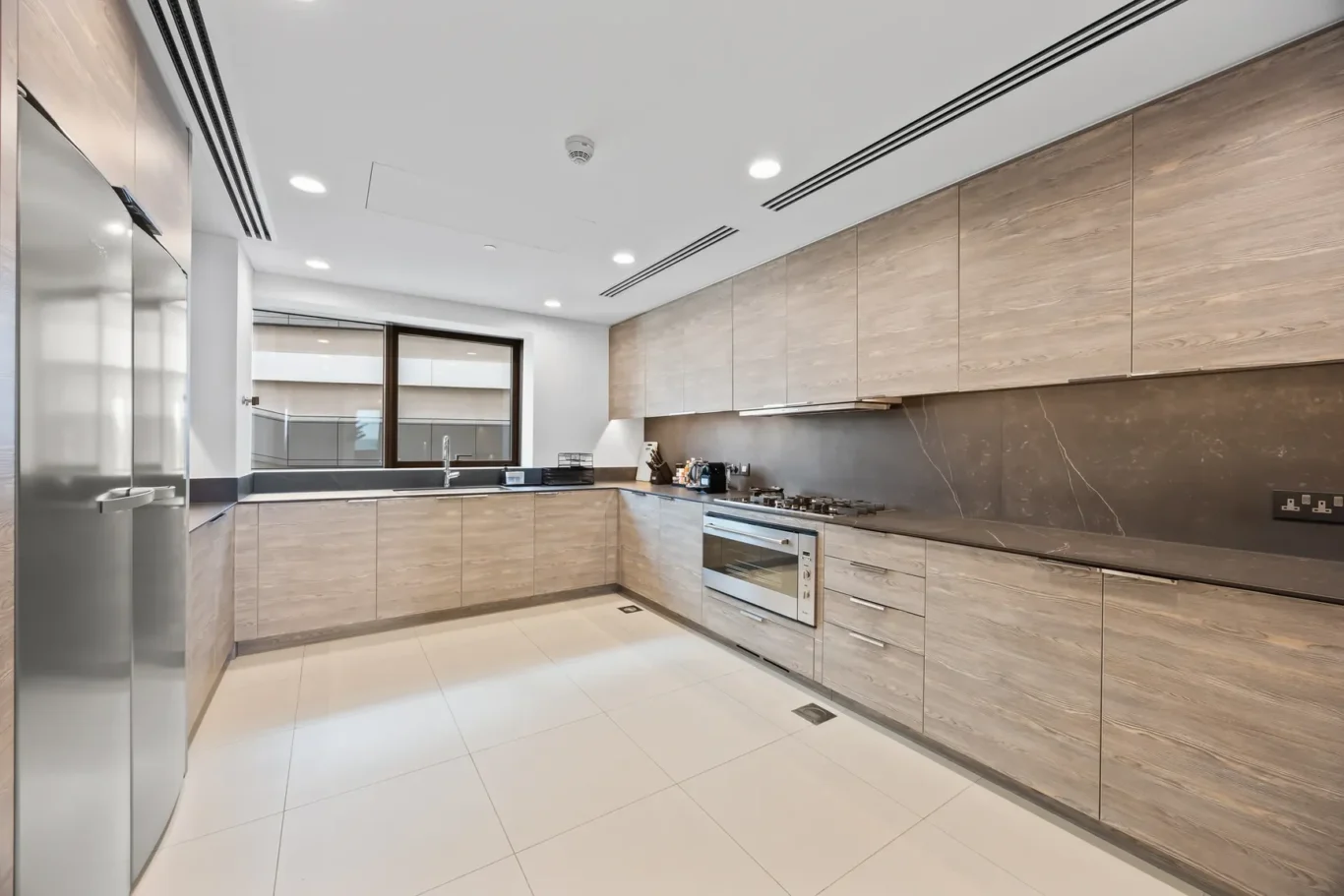 Modern L-shaped kitchen with light wood cabinets, dark marble backsplash, and stainless appliances including an oven and gas hob.