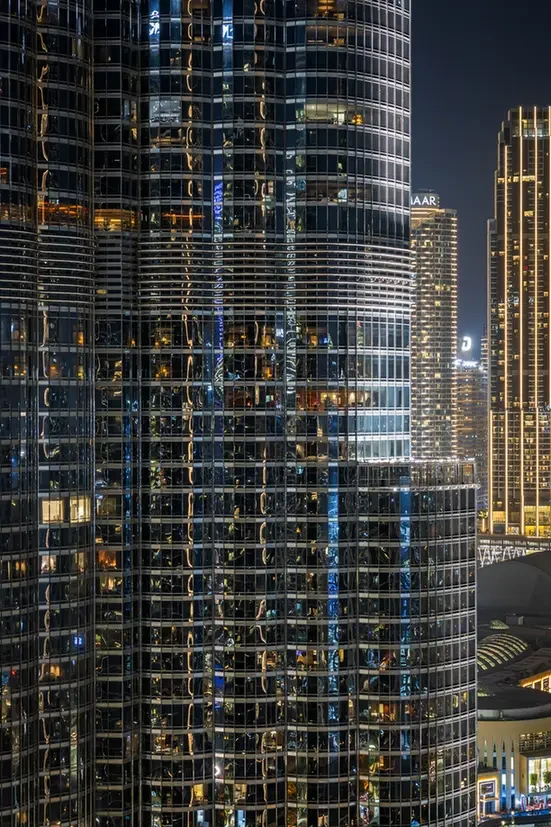 Nighttime cityscape with a curved glass skyscraper, its illuminated grid of windows reflecting lights from nearby buildings.