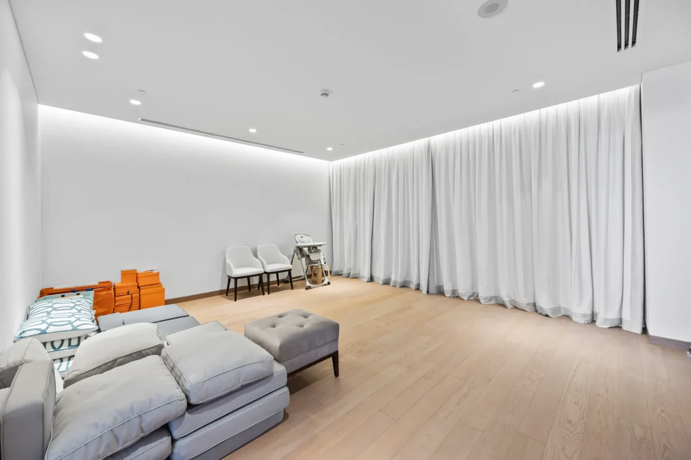 Bright minimalist room with a gray sectional sofa, orange storage boxes, two white chairs, a high chair, and tall white curtains along the wall.