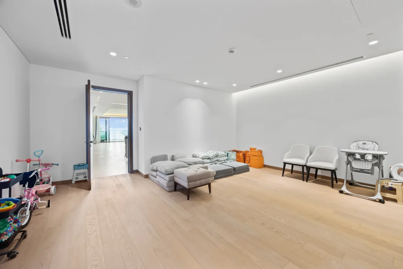 Spacious, modern living area with light wood floor, white walls, kids' toys on the left, a gray sectional in the center, and white chairs on the right; a high chair is at far right.