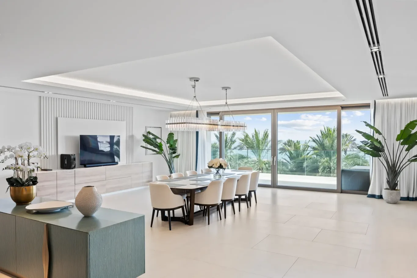 Bright modern dining room with a long table, cream chairs, and a chandelier, opening to a sunny patio garden through large glass doors.