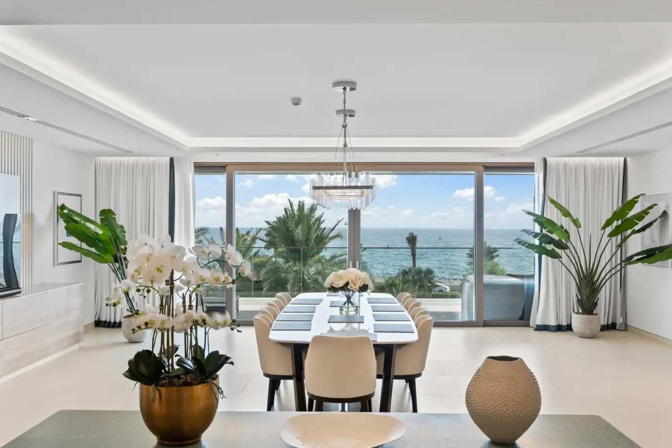 Bright coastal dining room with a long white table, beige chairs, and an ocean view through floor-to-ceiling windows.