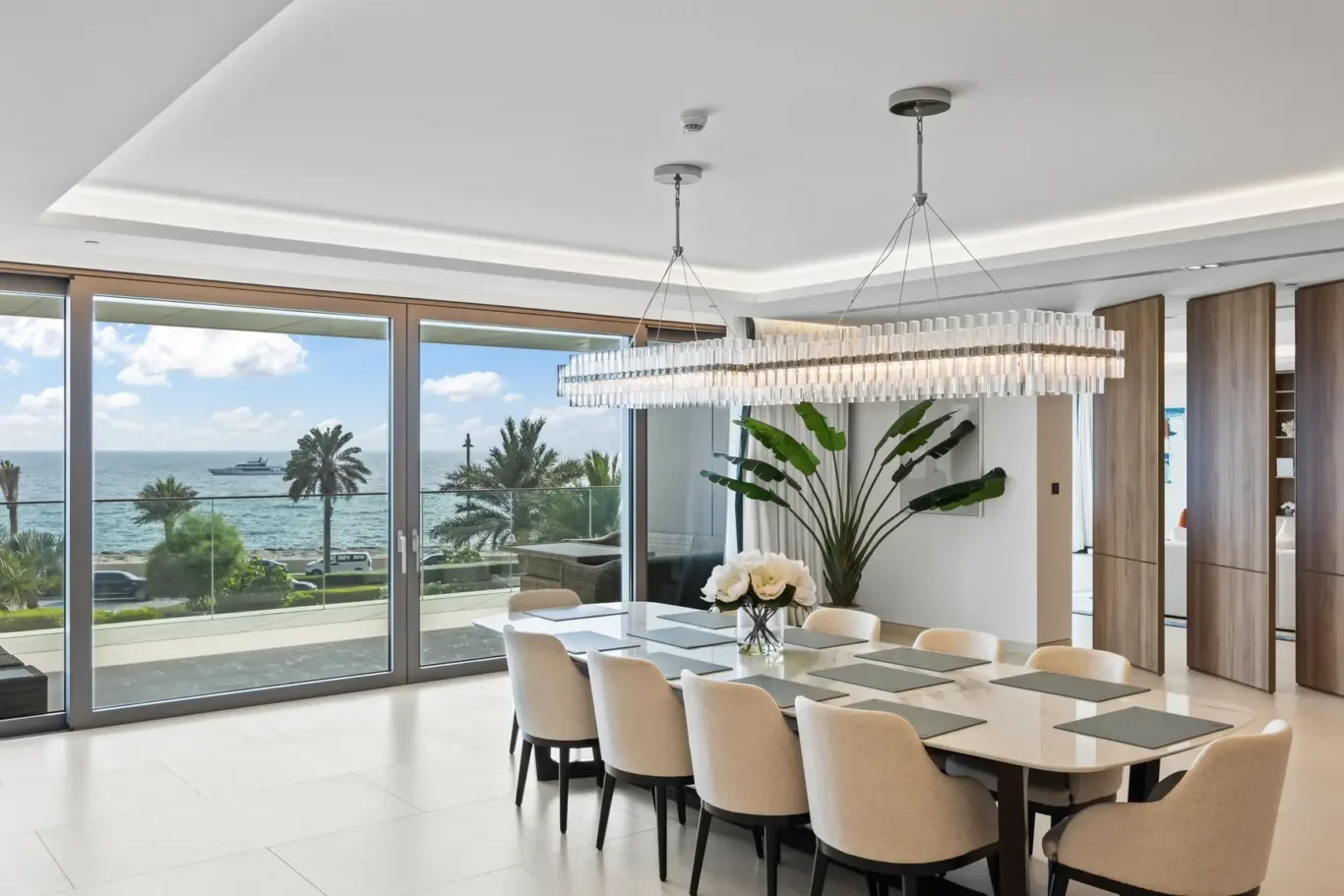 Modern coastal dining room with a long glass table and beige upholstered chairs, overlooking the sea through floor-to-ceiling glass doors; crystal chandelier hanging above.