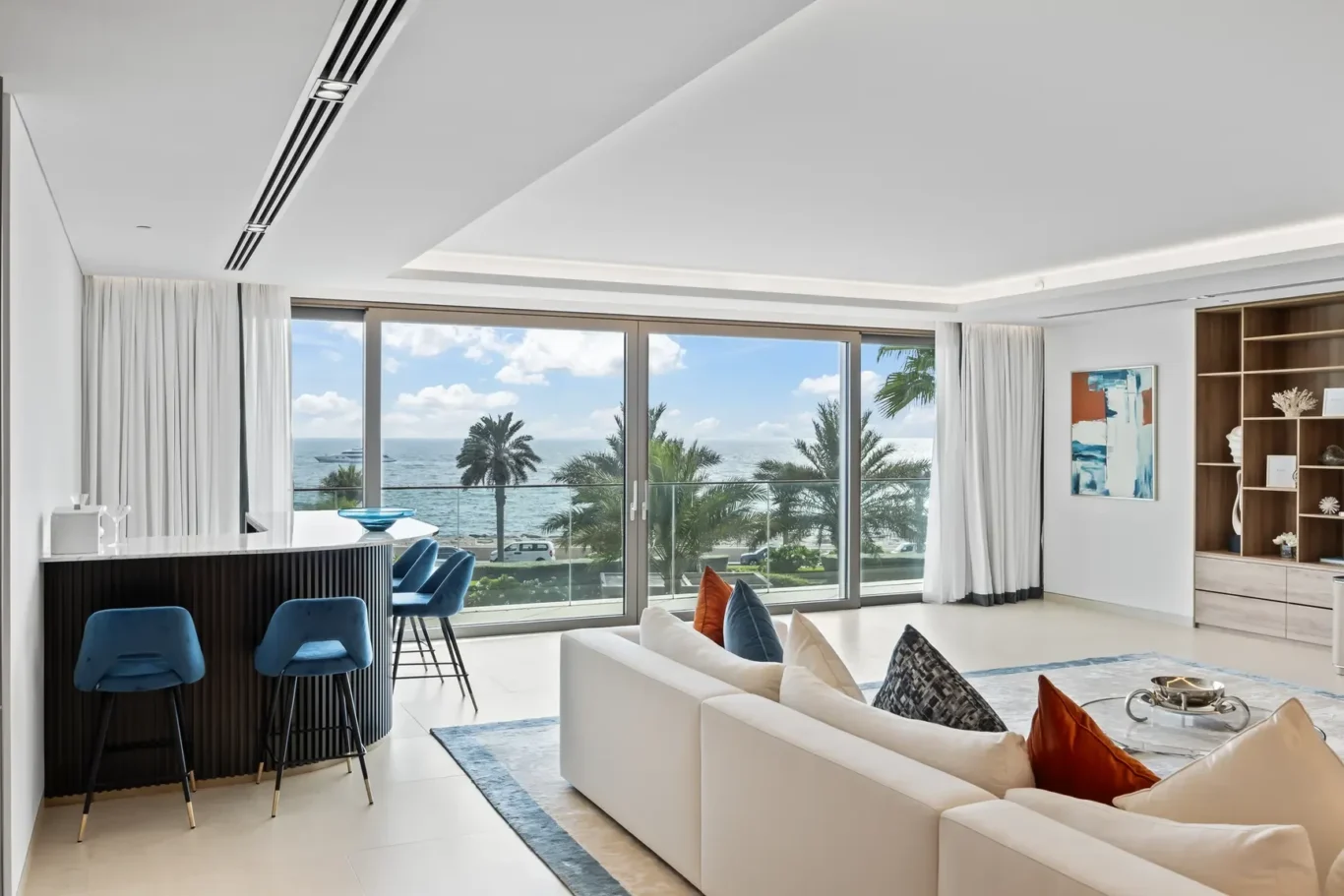 Bright coastal living room with white sofas, blue bar stools, and floor-to-ceiling windows overlooking a palm-lined ocean view.