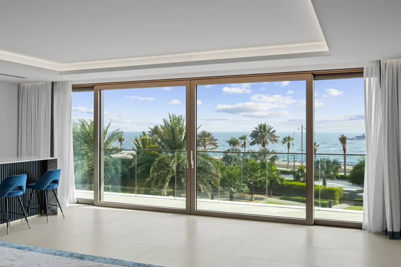 Modern living area with large wooden-framed sliding glass doors opening to a beach view of palm trees and the sea, white curtains on sides.