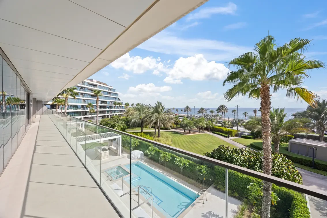 Seaside hotel balcony with glass railing overlooking a rectangular pool, green lawn, and palm trees against a blue sky and distant ocean.