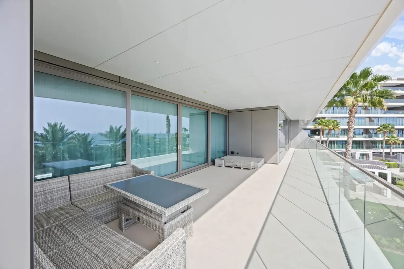 Spacious balcony with gray wicker seating, a glass-topped table, and a glass railing overlooking palm trees and a resort building.