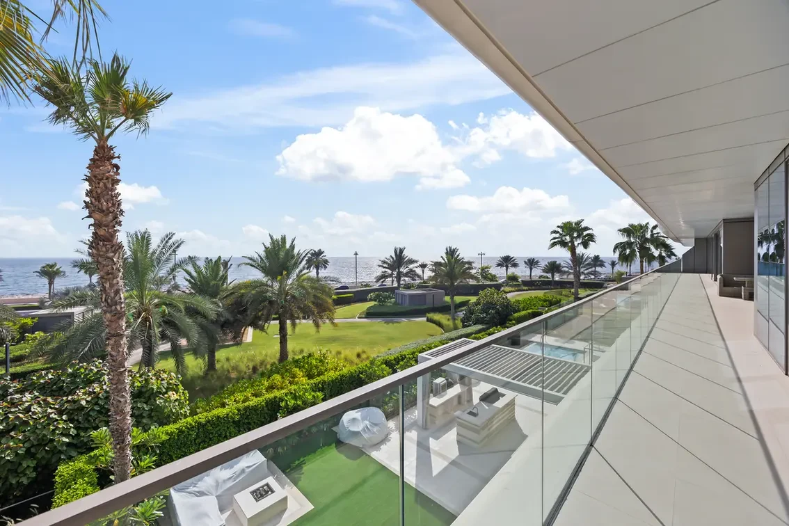 Oceanfront balcony with glass railing and white tiles, overlooking a palm-filled lawn and the sea under a blue sky with clouds.