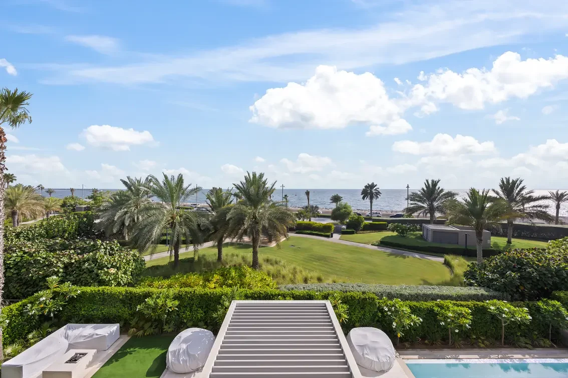 Coastal resort garden with palm trees, a manicured lawn, and the ocean visible in the distance under a blue sky.