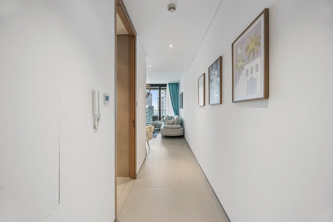 Bright, narrow hallway with white walls and framed art on the right, leading to a living area with teal curtains and a city view through large windows.