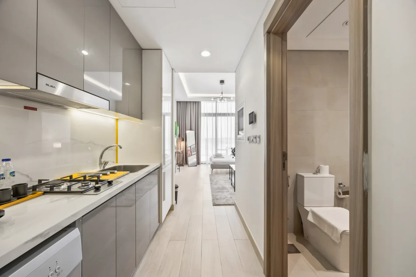Modern kitchen with gray glossy cabinets, white marble counters, gas stove and sink; open to living area with sofa and large windows.