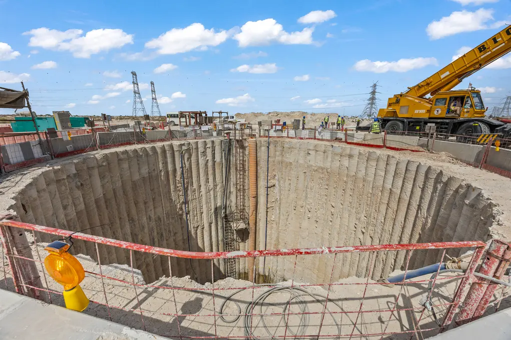 Construction site with a deep circular excavation pit and vertical concrete walls, an orange pipe along the pit, and a yellow crane on the right.