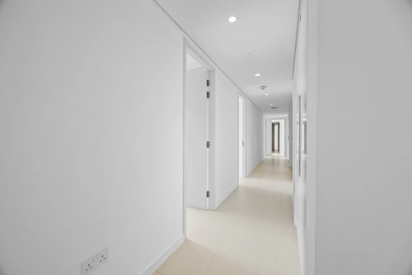 Long white hallway in a modern building with doors along both sides and ceiling lights clearly visible in perspective.
