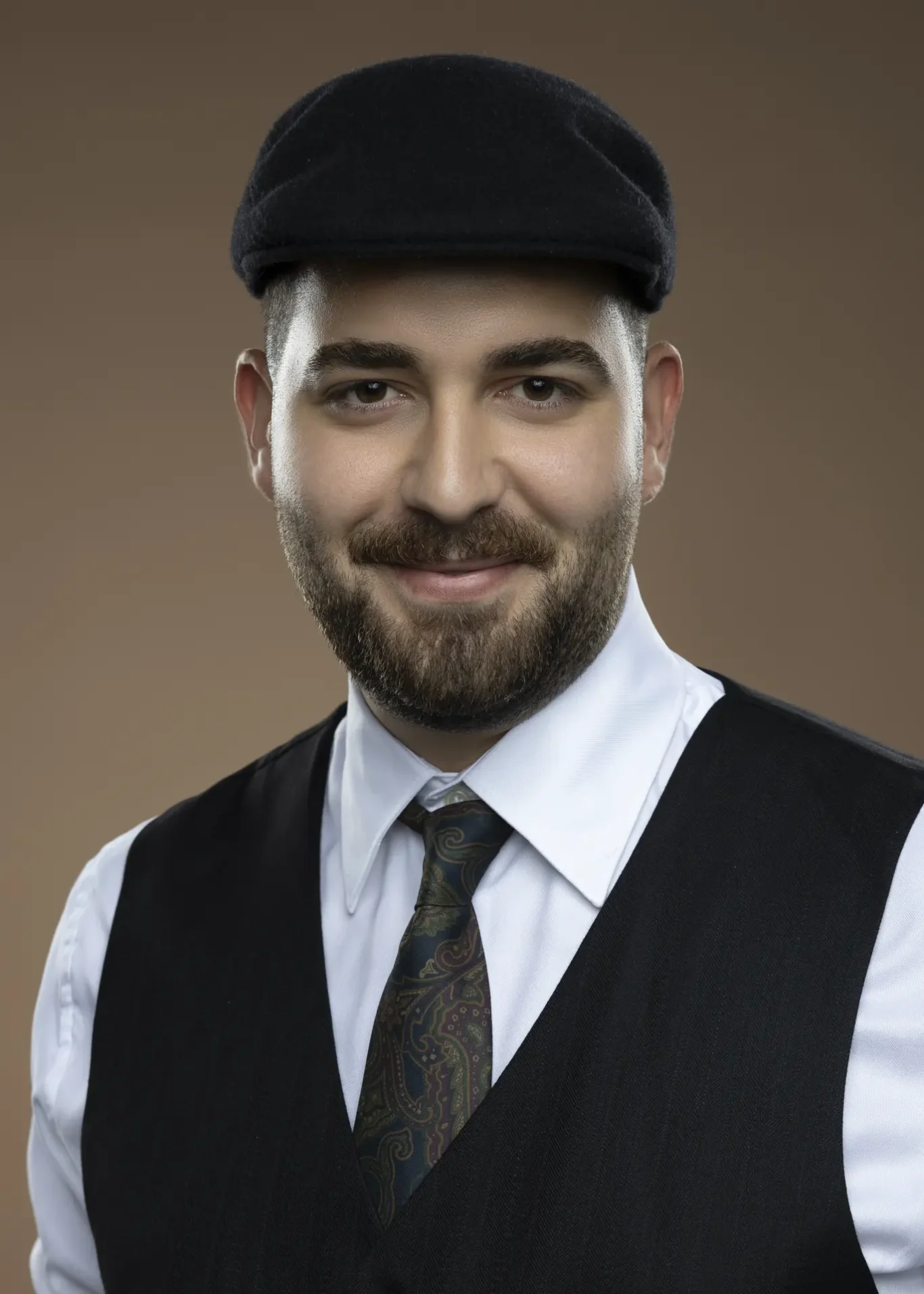 Smiling man in a black flat cap, white shirt, black vest, and paisley tie against a brown background.