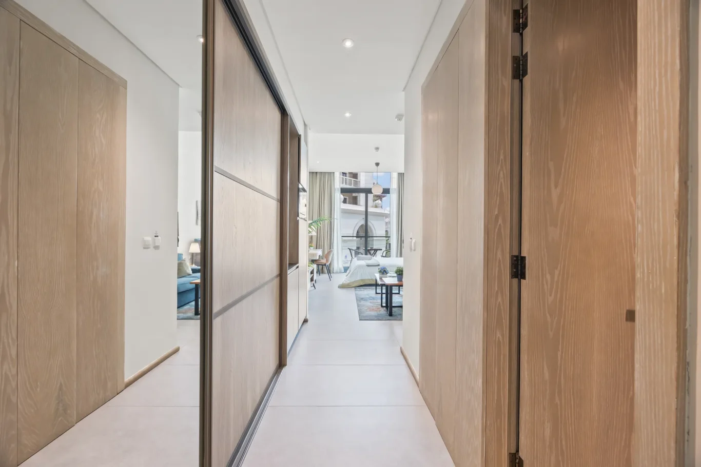 Long hallway with wood wardrobes on both sides and mirrored panels, opening to a bright living area with seating and a large window at the end.