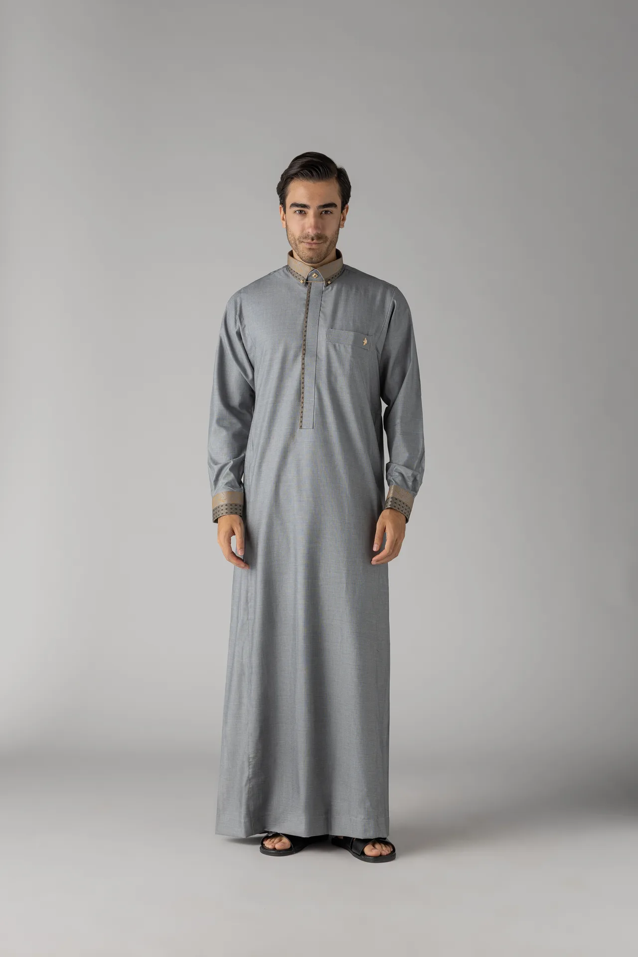 Man wearing a light gray traditional thobe with beige trim, standing against a plain gray backdrop.