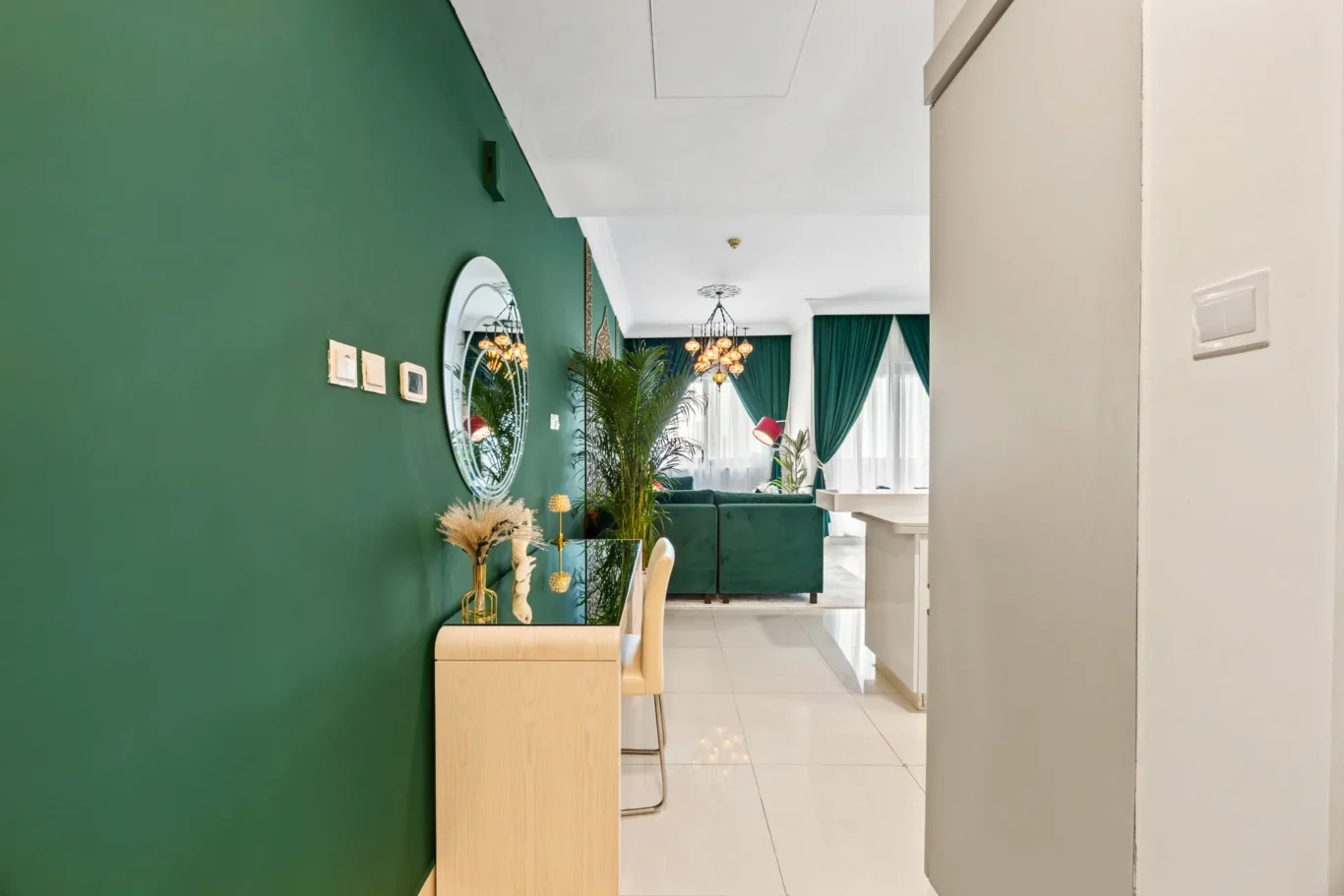 Indoor living space with a dark green accent wall, round mirror, and light wood console table with beige chairs nearby.
