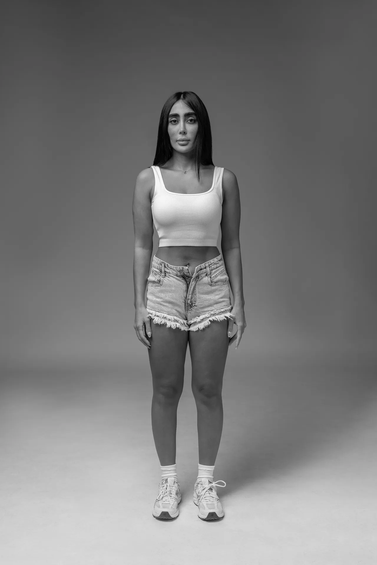 Full-body portrait of a young woman facing the camera, wearing a white tank top, frayed denim shorts, and sneakers in a grayscale studio setting.