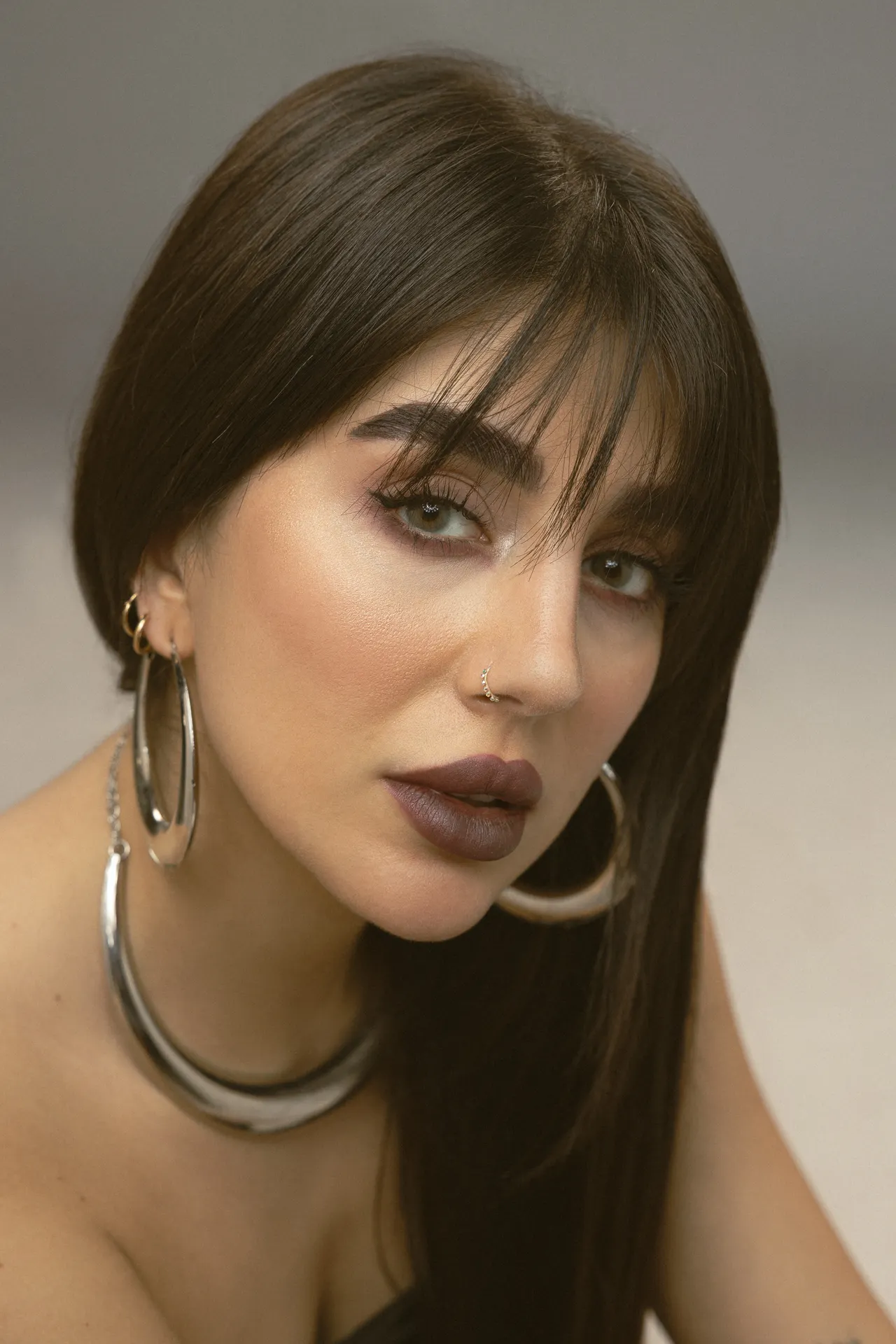 Close-up portrait of a woman with dark brown hair, smoky eye makeup, and matte brown lips, wearing large silver hoop earrings and a chunky necklace, looking slightly to the side.