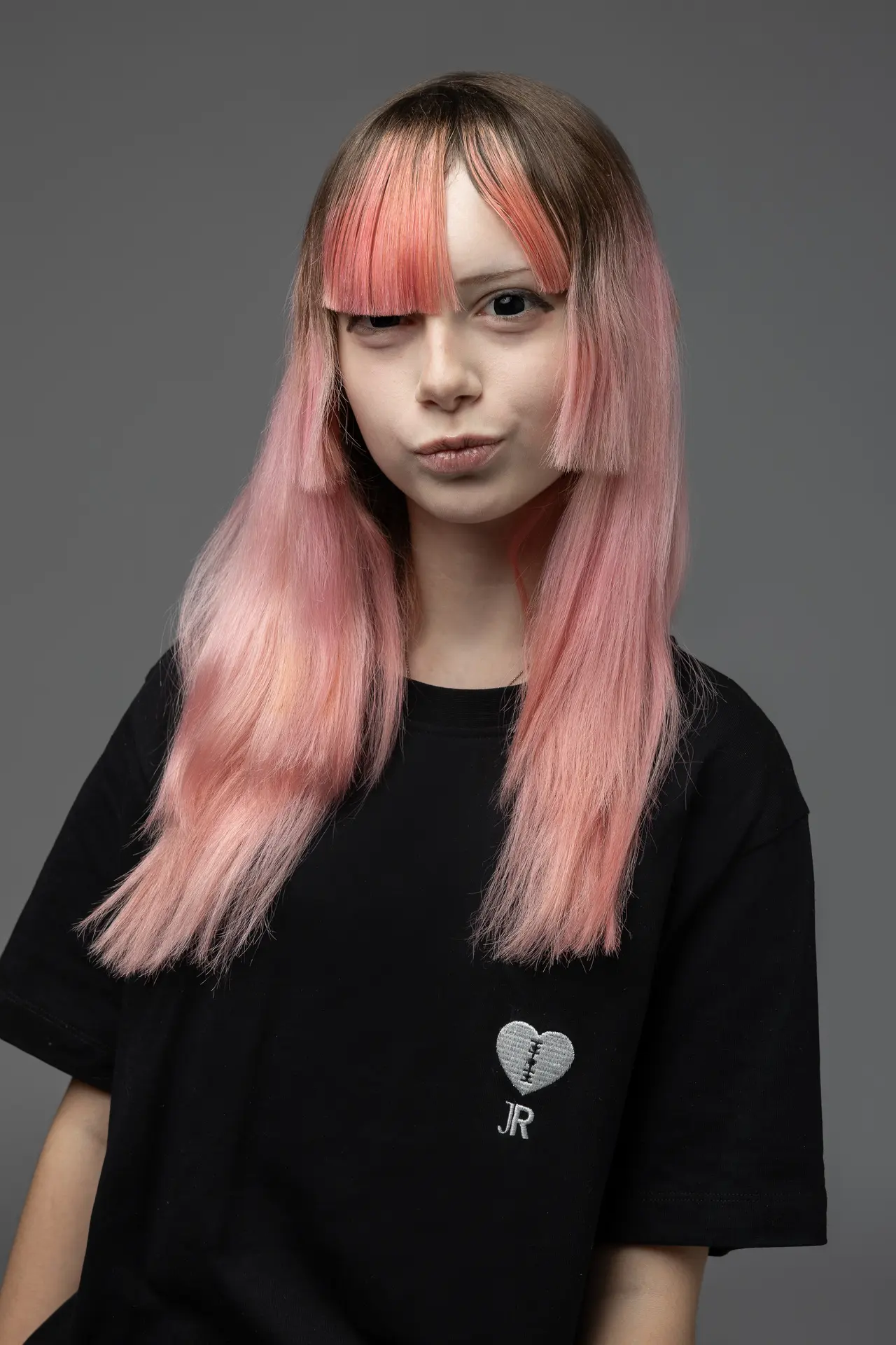 Portrait of a person with pink ombré hair and brown roots, wearing a black T-shirt with a stitched heart and 'JR' logo, against a gray background.