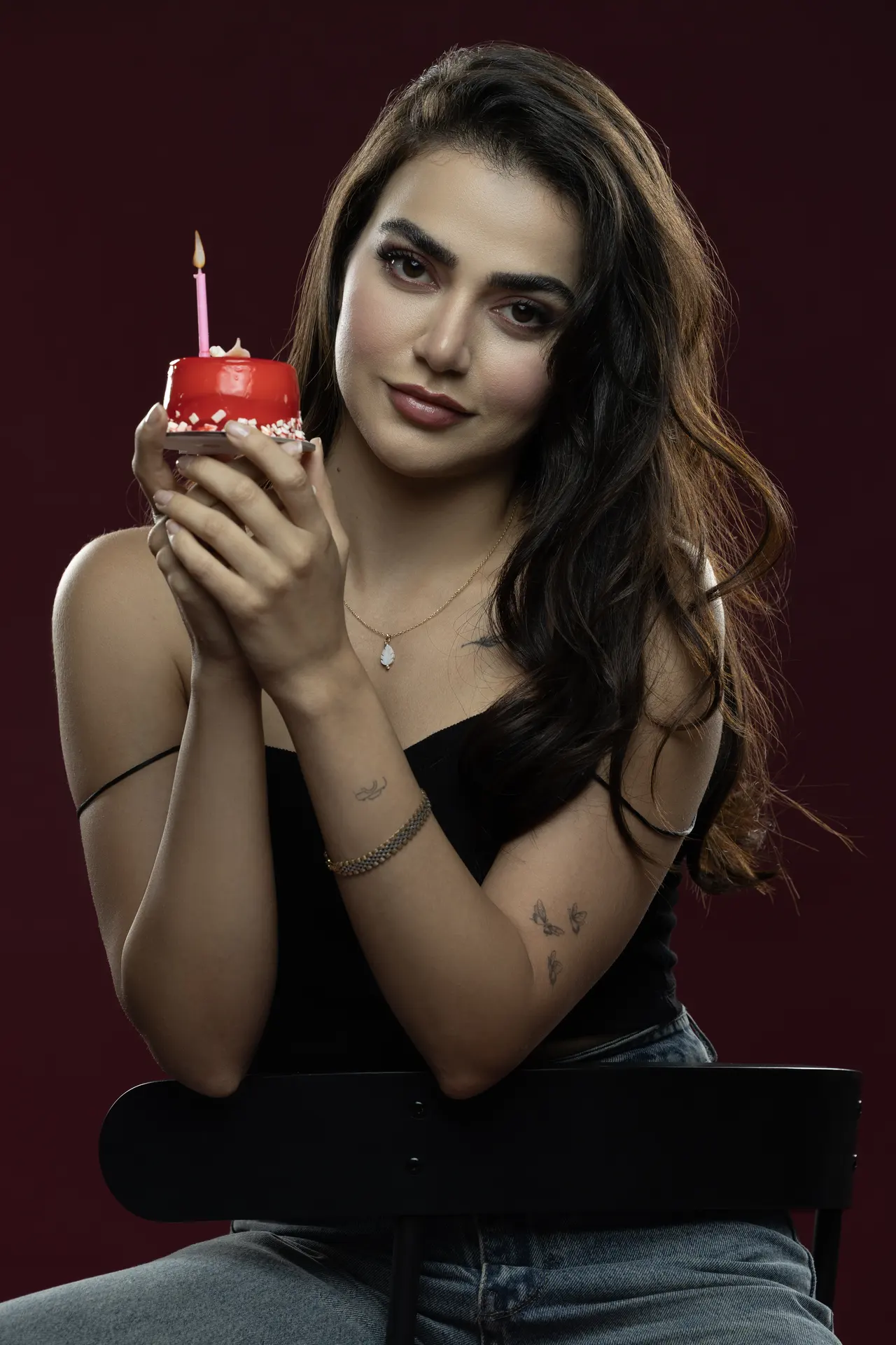 Young woman with dark hair holding a small red cake with a lit pink candle, posed against a dark maroon backdrop.