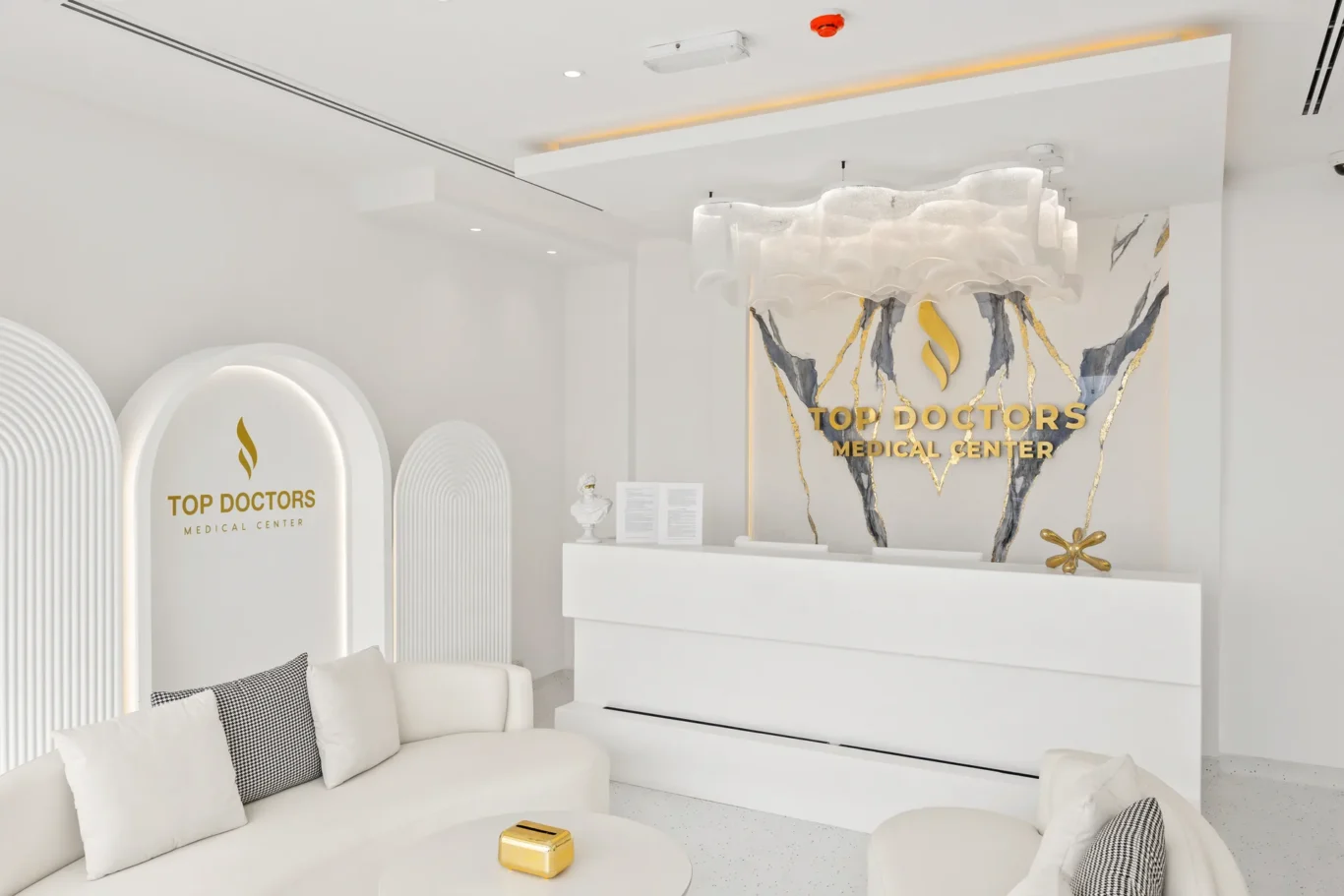 Reception lobby of Top Doctors Medical Center with white curved seating, round white tables, and gold-accented logo display on marble wall