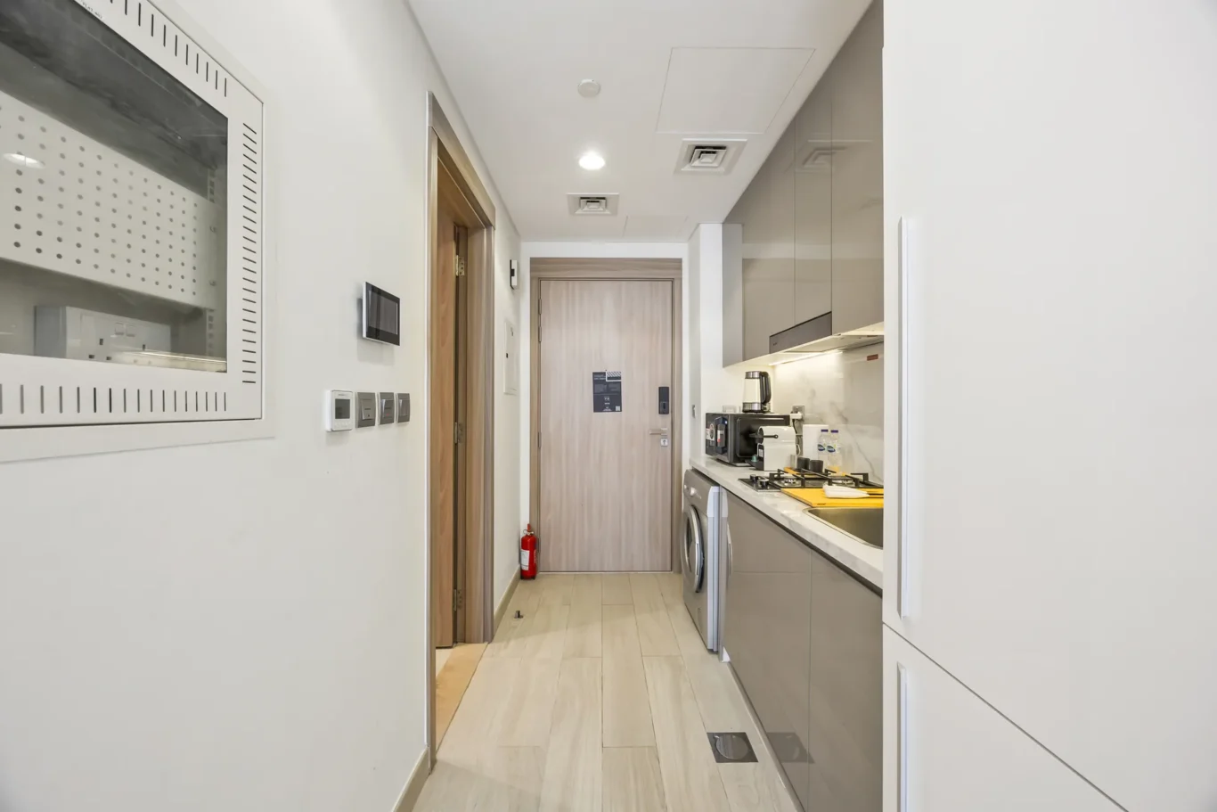 Modern galley kitchen along a hallway with grey glossy cabinets, washing machine, oven, kettle, and a wooden door at the end.