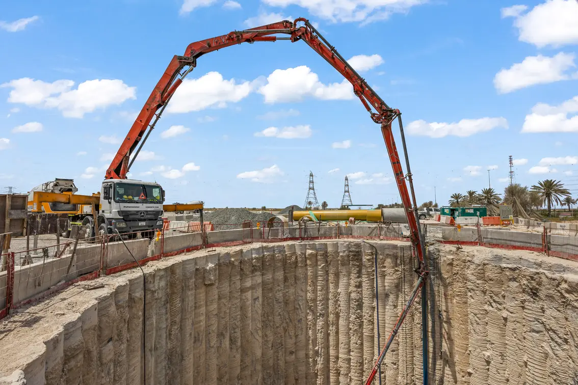 Construction site showing a red concrete pump truck arm arching over a deep circular trench, pouring into the pit with barriers around.