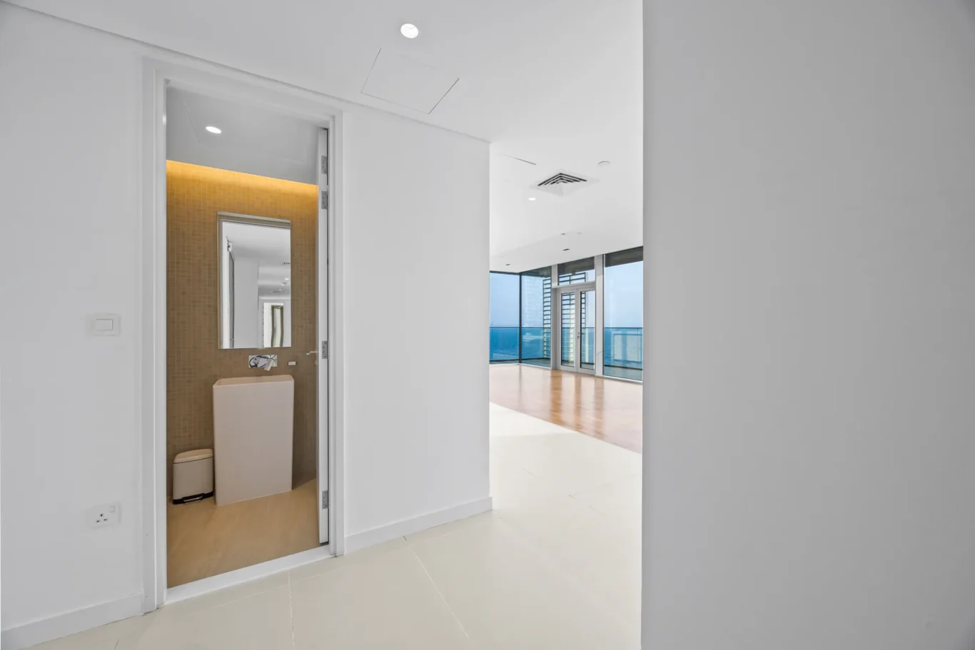 Open bathroom doorway with brown tiled wall, sink and mirror; bright living area with ocean view beyond.